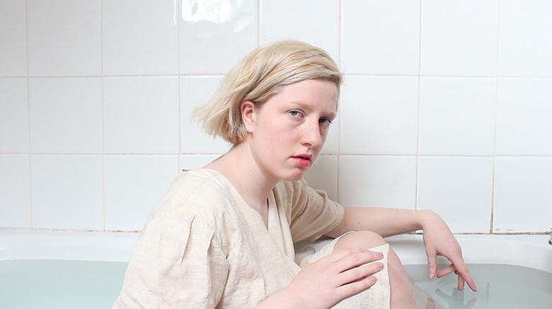 Still image from a video showing a young woman wearing white and seated in a bath full of water.