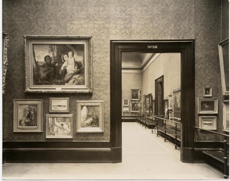 Black and white photo of paintings hung Salon-style on the wall with view through doorway into next gallery space