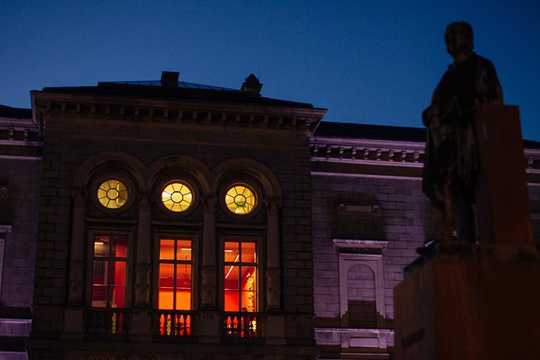 Nighttime photo of the exterior of the Merrion Square entrance to the National Gallery of Ireland with the windows illuminated and an outdoor sculpture silhouetted against the sky.