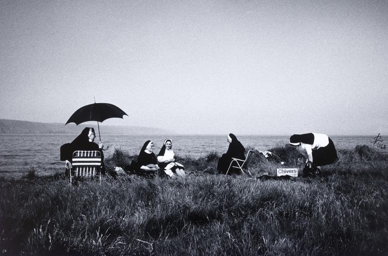 A group of nuns sit on a grassy patch overlooking the coast. Some sit on deckchairs, one with an umbrella over her head to shade her from the sun; others sit on blankets.