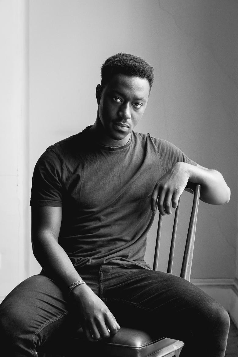 A black and white photograph of young man in jeans and a shirt sitting on a wooden chair, looking at the camera. One arm rests on the back of the chair.
