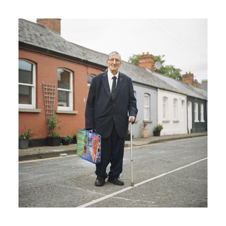 A man in a large, baggy black suit stands in front of a row of cottages. He is leaning on a crutch. In the other hand he carries a blue shopping bag.