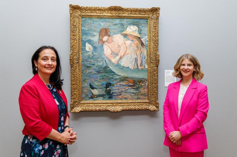 Photograph of two female figures, one wearing a bright pink suit and one wearing a navy dress with a red jacket, standing in front of a painting of two female figures in a boat