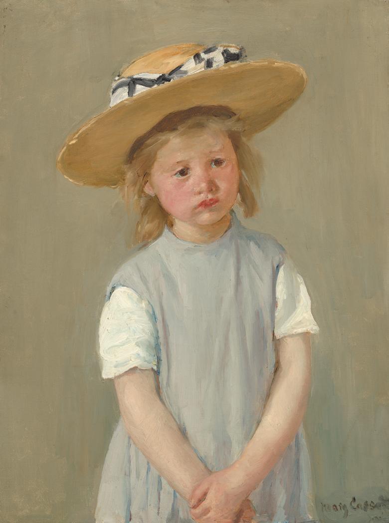 A child in a blue smock with white short sleeves and a large sunhat stands with her hands crossed in front of her. 