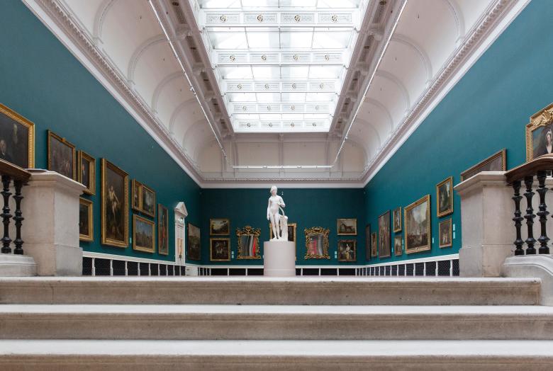 Large green room with high ceilings, a glass ceiling, walls hung with paintings and a white marble statue in the middle of the room.