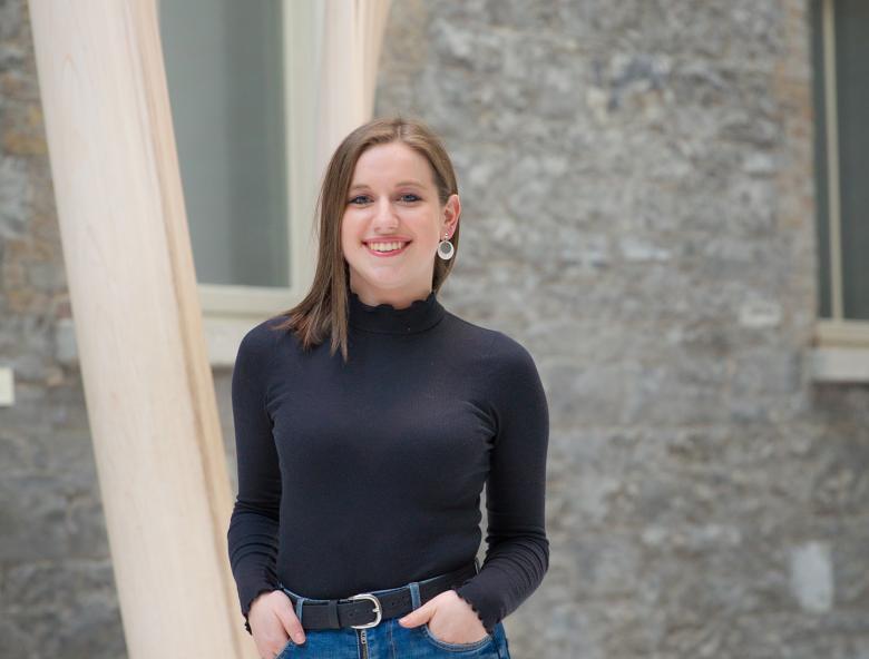 Sarah stands in the Gallery Courtyard. She is smiling, with her hands in her pockets.