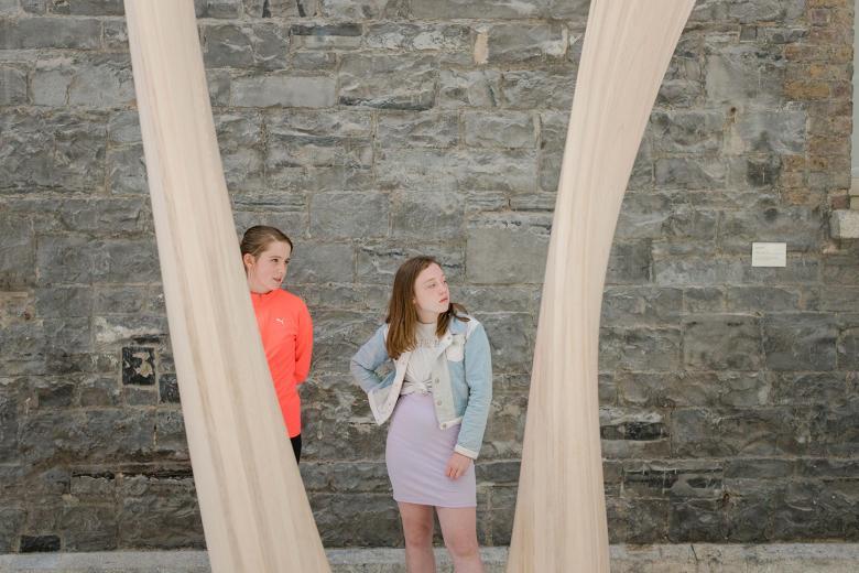 Two young girls standing in front of a stone wall framed by a curved wooden sculpture