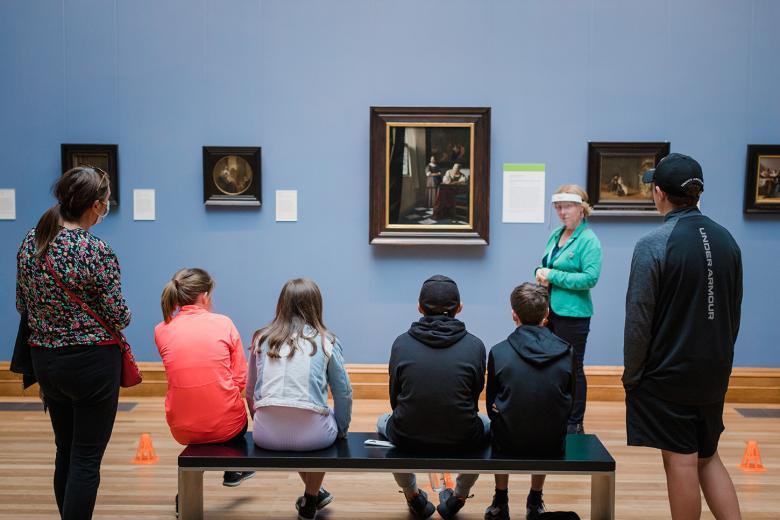 A woman wearing a face shield standing in front of Vermeer's Woman Writing a Letter with her Maid, speaking to a group of 5 school students and their teacher.