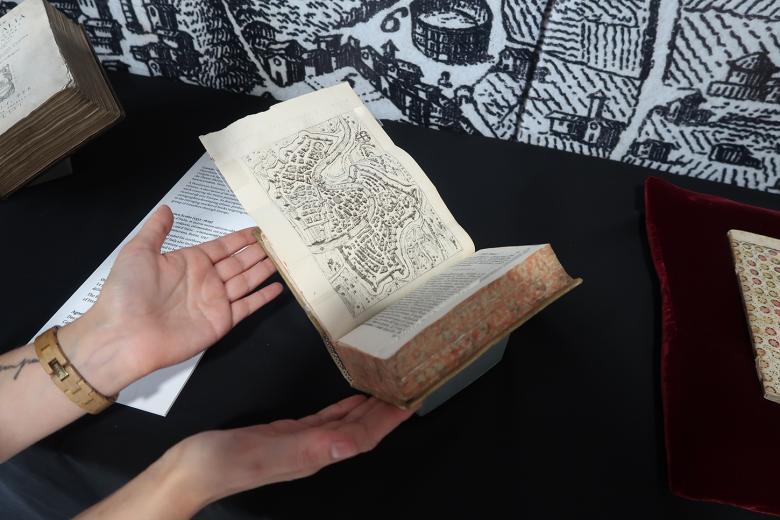 A rare travel book lies open on a black velvet cushion, showing a map. A pair of hands reach towards it.
