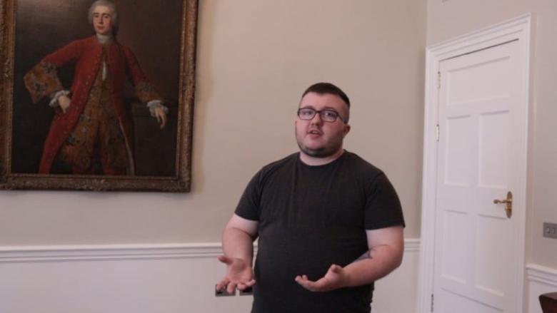 Video still of a young trans man standing in front of a gilt-framed portrait in a room with pale walls and a white door.