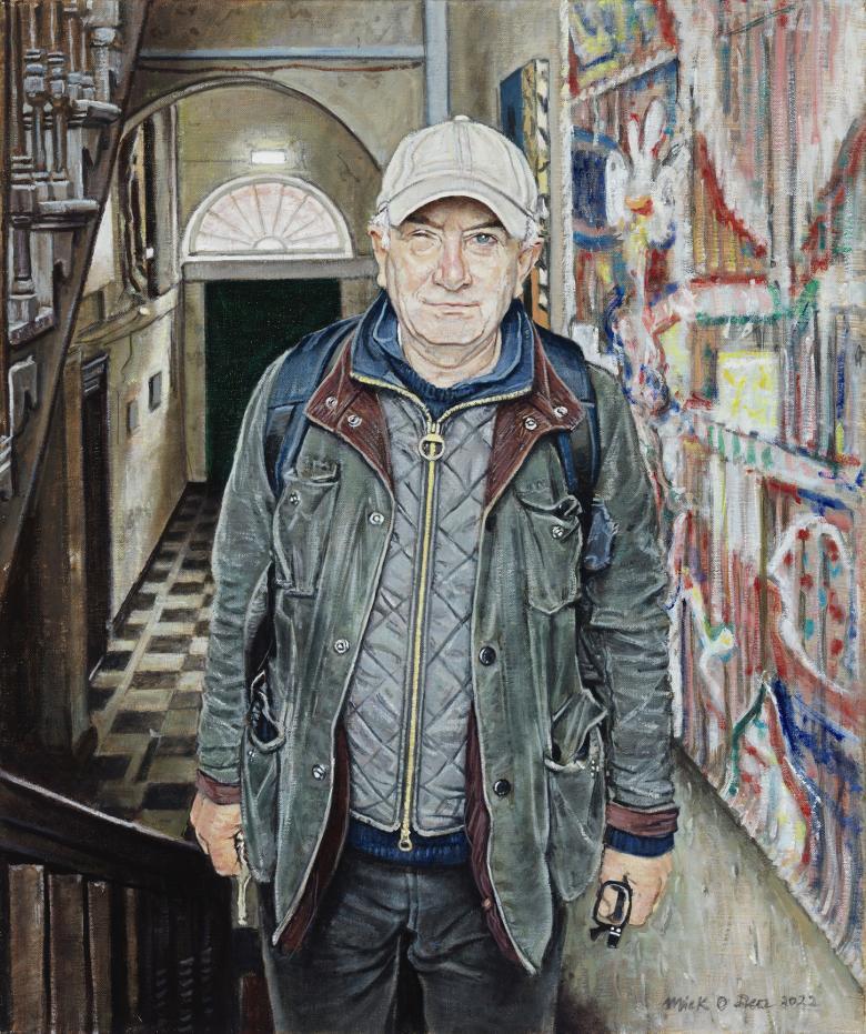 A man stands in a stairwell. Behind, we see a door with a fanlight and a tiled hallway. To his left, a colourful painting hangs on the wall. He is wearing jeans, a baseball cap, a wax jacket and a puffer jacket, and has a backpack on his back.