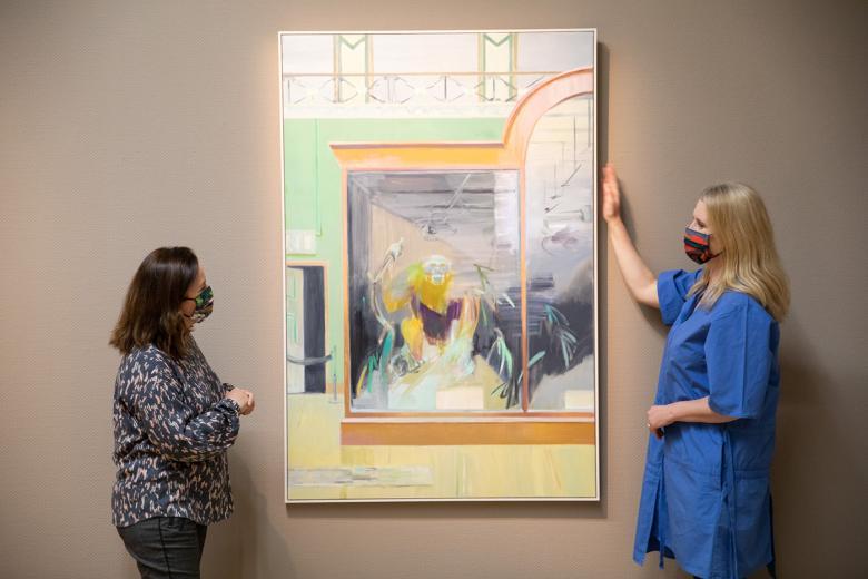 Two women wearing face masks standing either side of a painting that's hanging on a grey wall