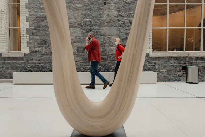 A man and woman wearing face masks walk past a wooden sculpture in the Gallery