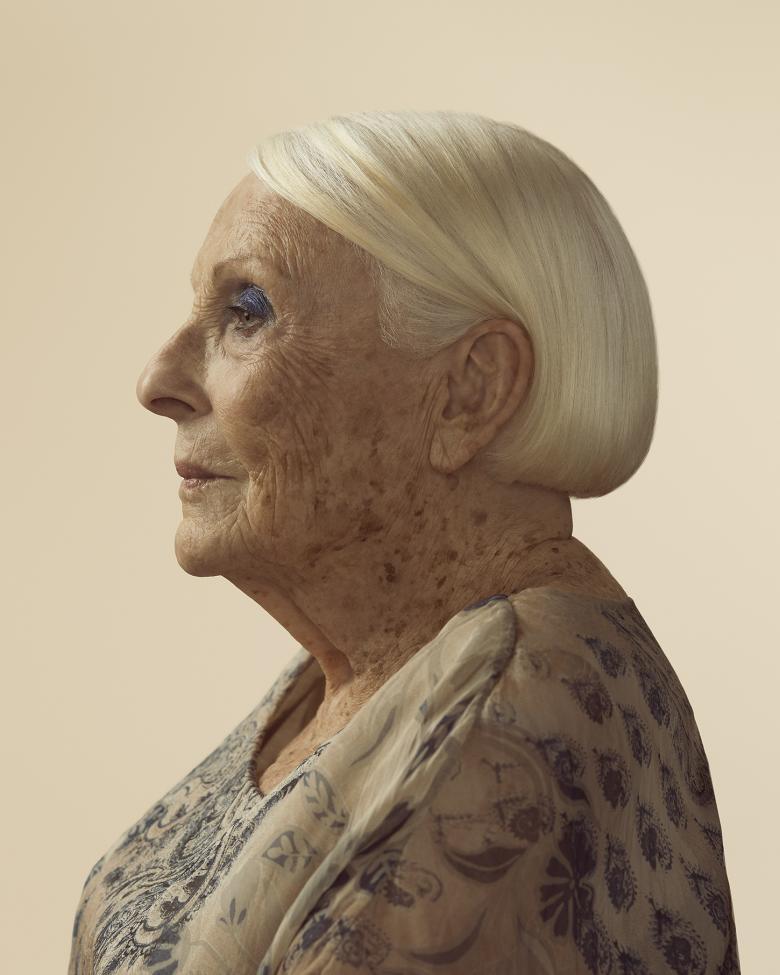 A photograph of a woman in side profile. She has a sleek white bob tucked behind her ear and wears dark blue eyehadow. She wears a white blouse with ink blue patterns on it.