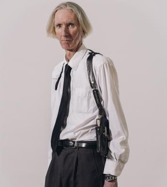 A three quarter length portrait of a male figure dressed in a white shirt, black tie, and back pants. He has white hair, and is looking directly at the viewer. He wears a black holster across his shoulders, which, when we look closely, holds a notebook and pen. 