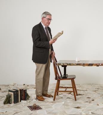A man stands to the left of the frame wearing a suit. He is reading from a book, and he rests his hand on a chair in front of him. There is another chair opposite, and a table rests on the seat of the chairs. Strewn around the floor are pages from a book, and to the left is a small selection of books propped up between bookends. To the right is an efigy of a harp which appears to be made from coins.