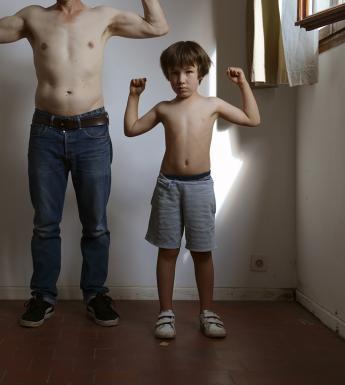Two figures - an adult and a child - stand in an empty room against a white wall. Both are topless, with the adult figure in jeans and the child in shorts. They both have their arms raised as though holding an imaginary weight over their heads. The adult figures head is not visible, but the child looks directly at the camera, and there is a shaft of light from the open window behind him.