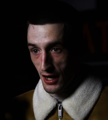 A face peers out of the shadows. The man wears a zipped up jacket with a white furry collar. His mouth is partially open, and his eyes look past the viewer, to the left of the frame.
