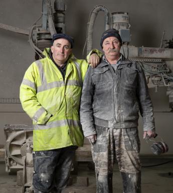 Two men stand in front of industrial-looking equipment. They wear work clothes and boots, and are covered in dust. The man on the left wears a high-vis jacket and rests his hand on the shoulder of the second man.