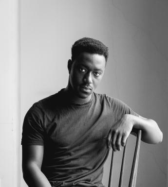 A black and white photograph of young man in jeans and a shirt sitting on a wooden chair, looking at the camera. One arm rests on the back of the chair.