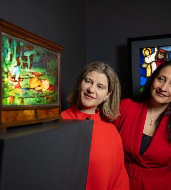 Photograph of two women, both wearing red dresses, looking around a corner at a piece of decorative stained glass.
