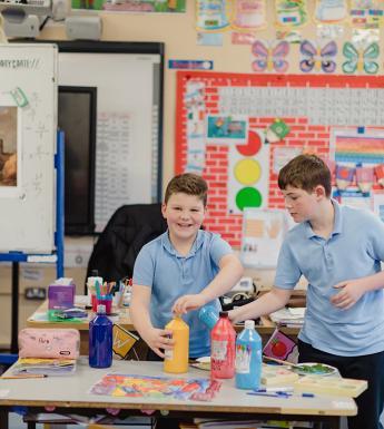 Two boys in blue polo shirts stand behind a table covered in art supplies, smiling at the camera