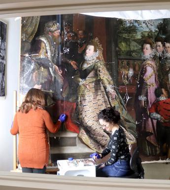 View through a window into a conservation studio where two women work on a large oil painting