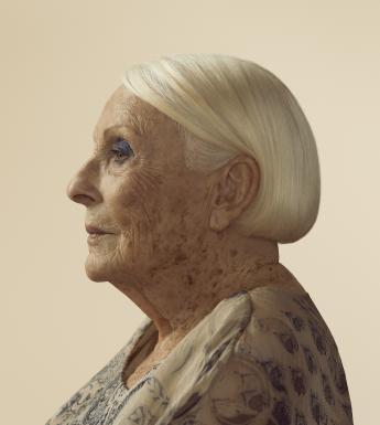 A photograph of a woman in side profile. She has a sleek white bob tucked behind her ear and wears dark blue eyehadow. She wears a white blouse with ink blue patterns on it.