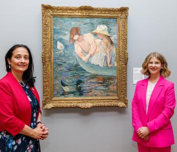 Photograph of two female figures, one wearing a bright pink suit and one wearing a navy dress with a red jacket, standing in front of a painting of two female figures in a boat